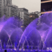Illuminated fountain in Macau with purple and blue water jets arching across a city plaza at dusk, framed by high-rise buildings, trees, and a decorative street lamp.