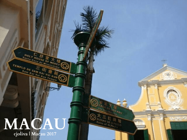 A green directional signpost in Macau pointing to the Holy House of Mercy, Sam Kai Vui Kun Temple, Lou Kau Mansion, and Ruins of St. Paul, with a colonial yellow church in the background under sunny skies.