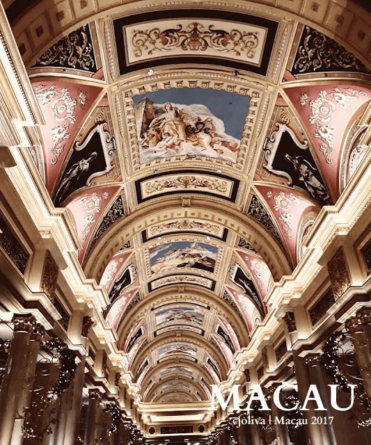 Ornately decorated interior ceiling in Macau featuring grand arches, elaborate frescoes, gold and red architectural details, and illuminated columns with festive decorations.