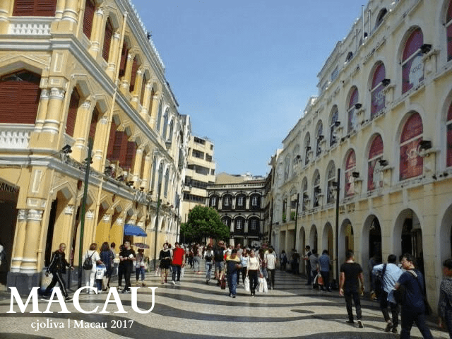 Bustling pedestrian street in Macau with pastel colonial-style buildings featuring arched windows and decorative facades, wave-patterned mosaic pavement, and crowds of people under a clear blue sky.