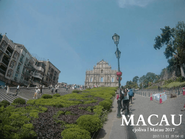 Ruins of St. Paul in Macau with a 17th-century Portuguese church facade, wide staircase, trimmed garden bushes, colonial buildings, and tourists walking and taking photos.