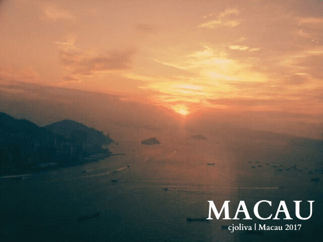 Sunset view in Macau, with boats scattered across the water, a warm orange-and-pink sky, clouds partially obscuring the sun, and mountainous islands in the background.