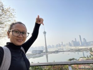 Traveler on a viewing platform in Macau, pointing toward the Macau Tower with the city skyline, bridge, and water under a clear blue sky.