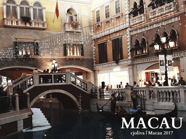 Indoor Venetian-style canal in Macau with a gondolier steering a gondola under a decorative bridge, surrounded by ornate Italian-inspired architecture, string lights, and people walking along the canal.