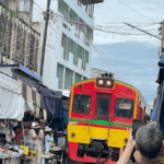 A bustling Thai market located directly on train tracks, with a slow-moving train passing through as vendors pull back their awnings and colorful umbrellas.