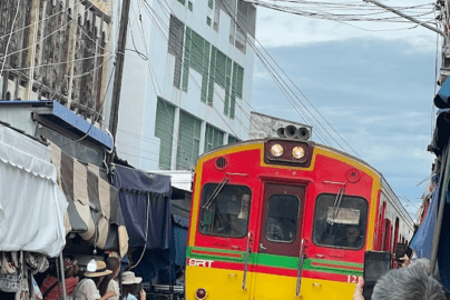 A bustling Thai market located directly on train tracks, with a slow-moving train passing through as vendors pull back their awnings and colorful umbrellas.