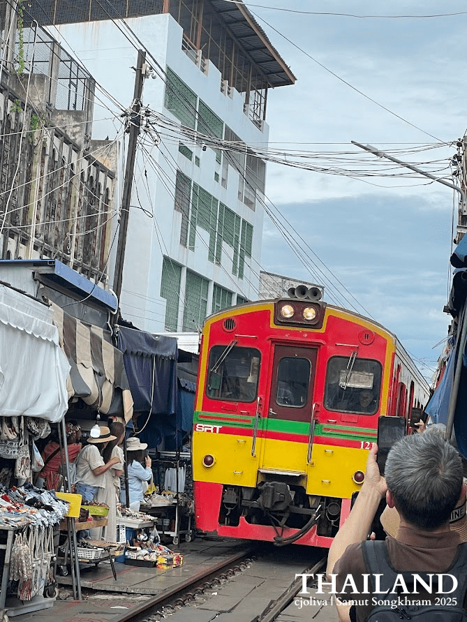 A bustling Thai market located directly on train tracks, with a slow-moving train passing through as vendors pull back their awnings and colorful umbrellas.
