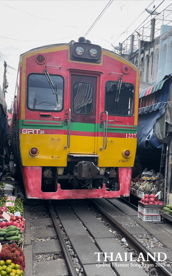 A head-on view of a red and yellow State Railway of Thailand train (No. 1227) passing extremely close to baskets of dragon fruit and tropical produce at the Maeklong Railway Market.