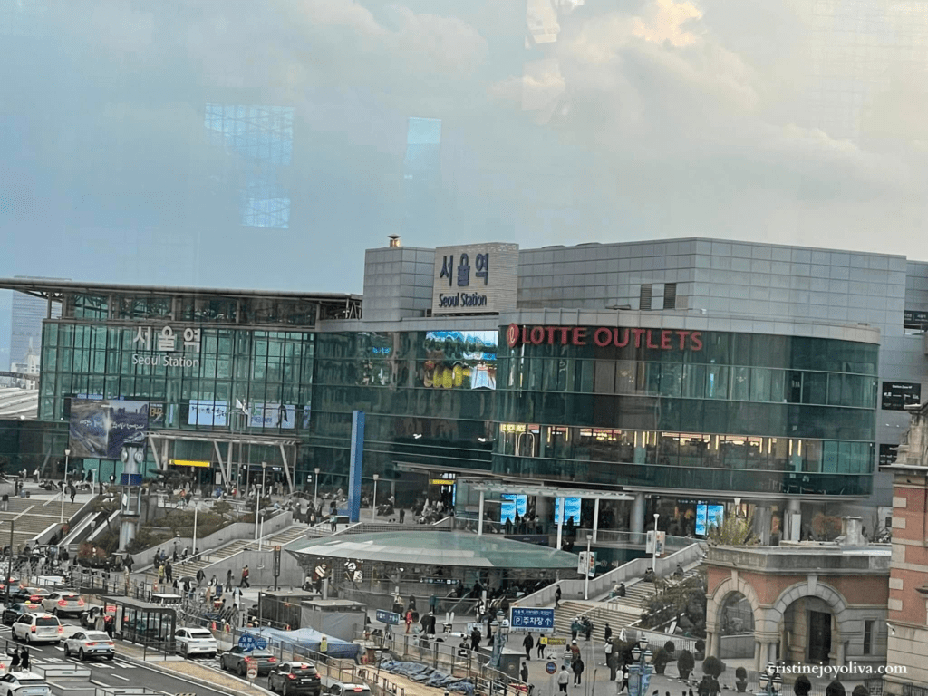 A high-angle view of the modern Seoul Station glass building and Lotte Outlets during a cloudy day, with busy city traffic and pedestrians in the foreground.