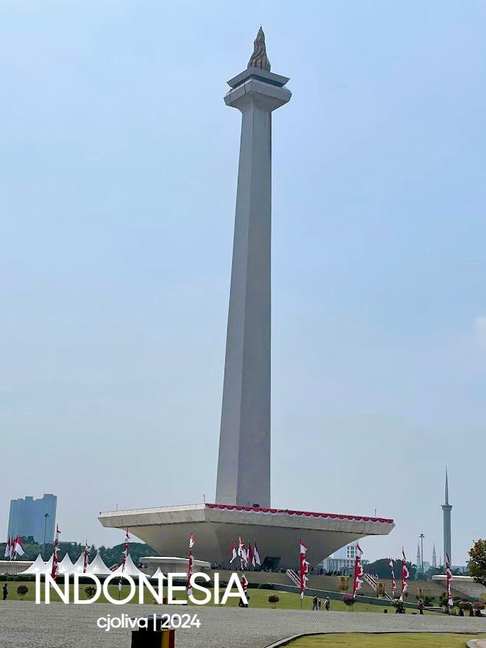 The National Monument (Monas), a tall white obelisk with a gold-leaf flame at the top, standing against a clear sky in Merdeka Square, Jakarta, surrounded by Indonesian flags.