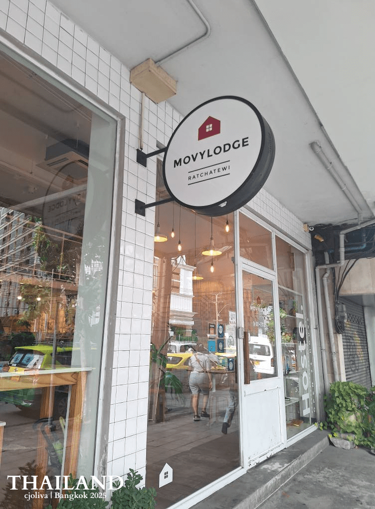 Exterior view of MovyLodge Ratchathewi in Bangkok, featuring a white tiled storefront, a circular hanging sign, and glass doors with hanging Edison bulb lights.