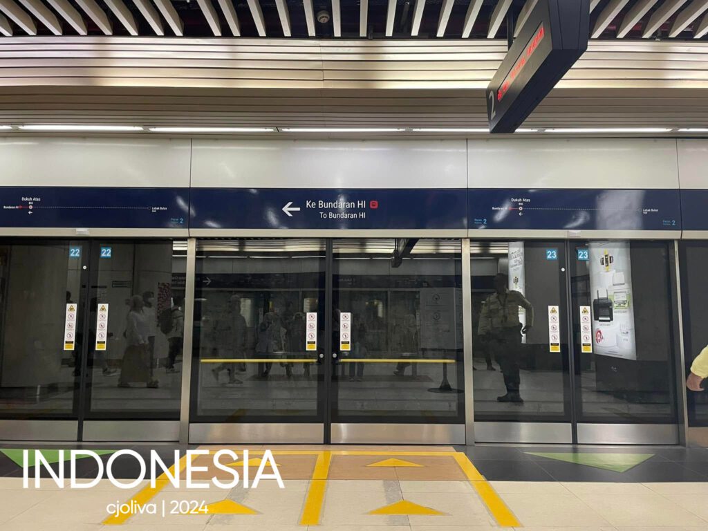 A modern underground subway platform in Jakarta, Indonesia, showing glass security doors with the sign "Ke Bundaran HI" (To Bundaran HI) and yellow directional floor markings for passengers.