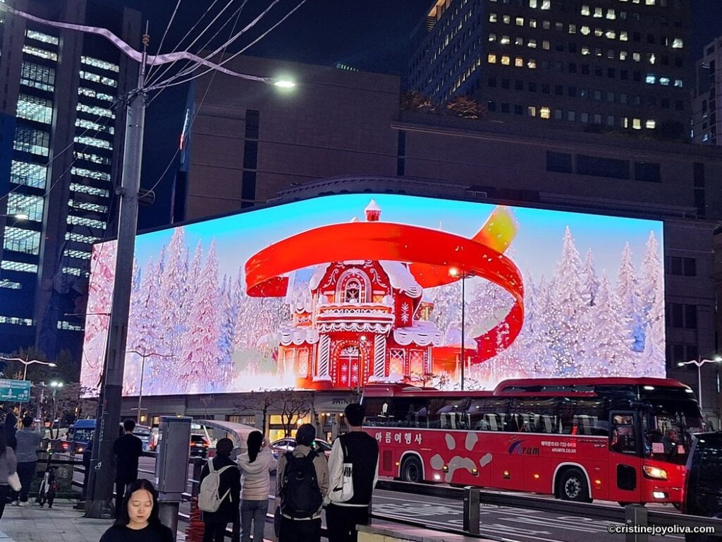 A festive 3D digital billboard in Myeong-dong, Seoul, at night, showing a gingerbread house with snow-covered trees and ribbon animation, with pedestrians and a tour bus nearby.