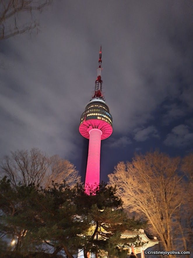 N Seoul Tower glowing with vibrant pink lights against the night sky, framed by silhouettes of trees and a traditional Korean pavilion.