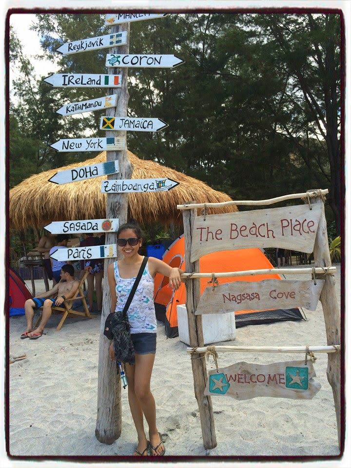 A traveler posing next to a rustic wooden signpost pointing to global cities like Paris and New York, located on the beach at Nagsasa Cove, Zambales.