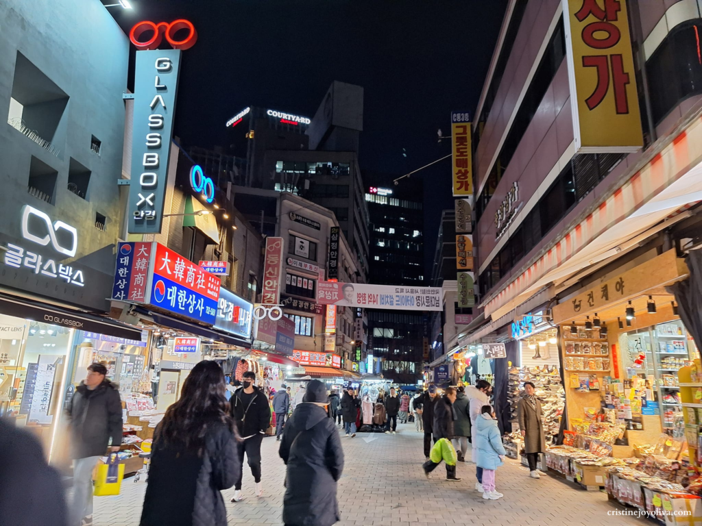 A busy street scene at Namdaemun Market at night, featuring bright neon shop signs, pedestrians walking through the market, and modern buildings in the background.