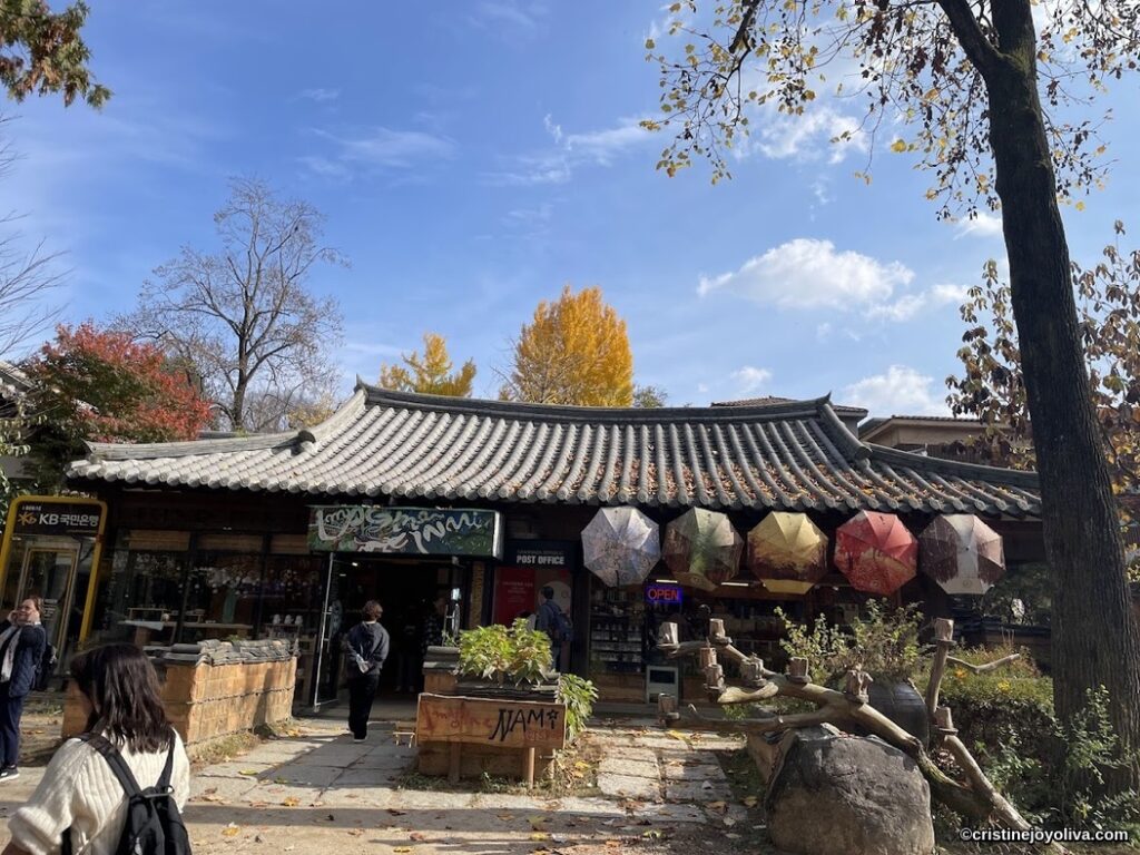 Traditional Korean-style building on Nami Island with tiled roof, autumn foliage, and colorful umbrella decorations above the entrance.