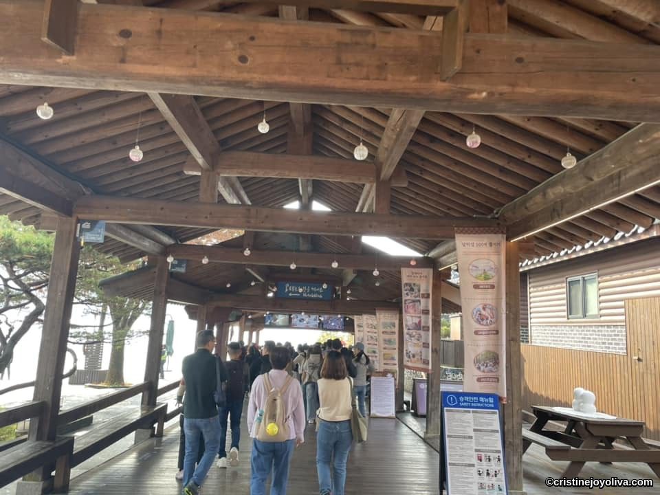 Visitors walking through a traditional wooden pavilion on Nami Island, South Korea, with hanging lights, banners, and surrounding trees.