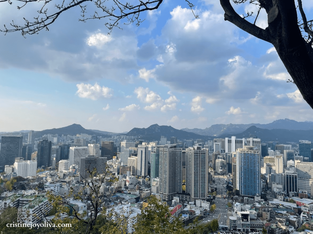 Panoramic view of the Seoul city skyline with modern skyscrapers and distant mountains under a blue sky, taken from a lookout on Namsan Mountain.