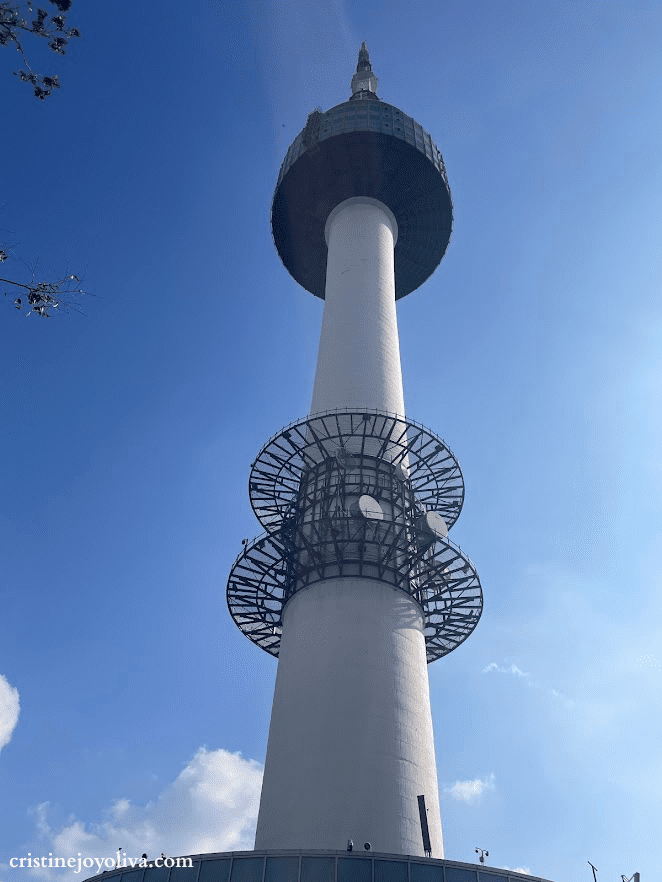 A dramatic upward-angle view of the N Seoul Tower against a clear blue sky, highlighting its white pillar and circular observation decks.