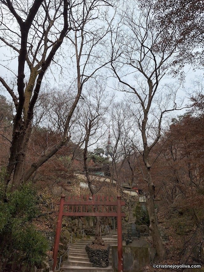 Stone steps on Namsan Trail in Seoul leading through a red torii gate toward N-Seoul Tower, framed by leafless trees and forest scenery.