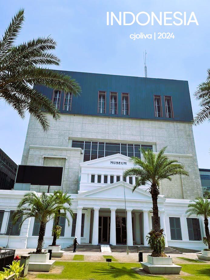 The white neoclassical facade of the National Museum of Indonesia in Jakarta, featuring grand columns, surrounded by palm trees and a larger modern building extension behind it.