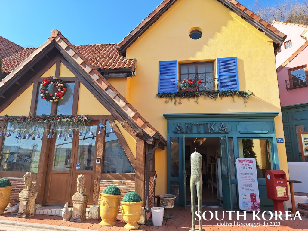 Colorful European-style street in Petite France, Gyeonggi-do, South Korea, featuring the Antika shop, a red mailbox, festive decorations, and a mannequin sculpture.
