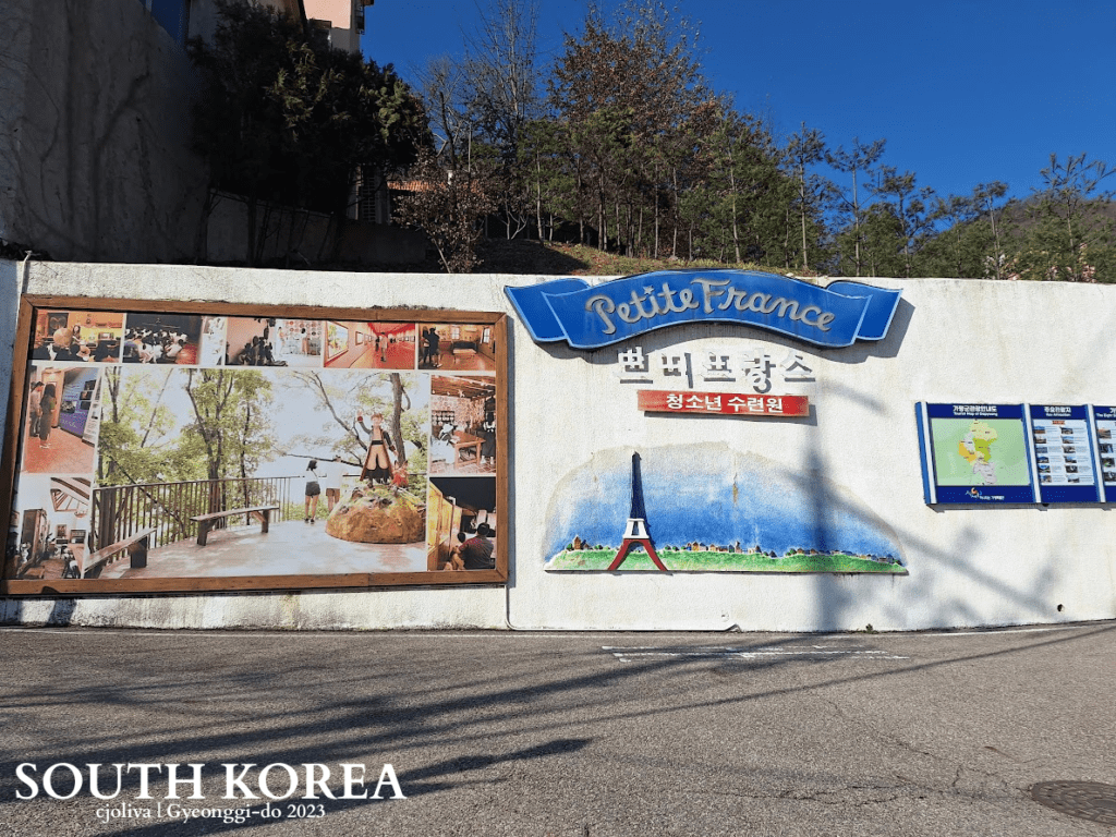 Entrance wall of Petite France cultural village in Gyeonggi-do, South Korea, featuring signage, Eiffel Tower mural, and visitor information boards.