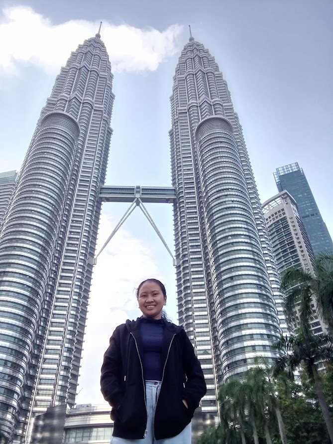 Traveler standing in front of the Petronas Twin Towers in Kuala Lumpur, Malaysia, with palm trees and a clear sky highlighting the iconic skyscrapers and skybridge.