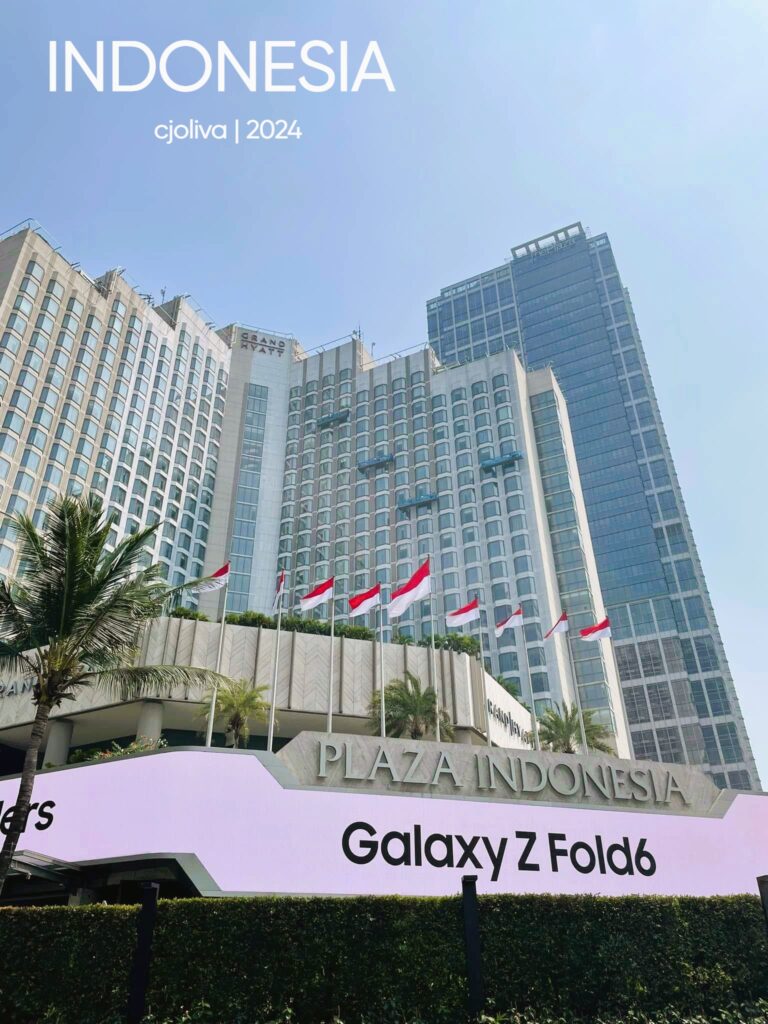 A daytime view of the Plaza Indonesia shopping mall and Grand Hyatt hotel building in Jakarta, with several Indonesian flags flying in front and a large digital billboard in the foreground.