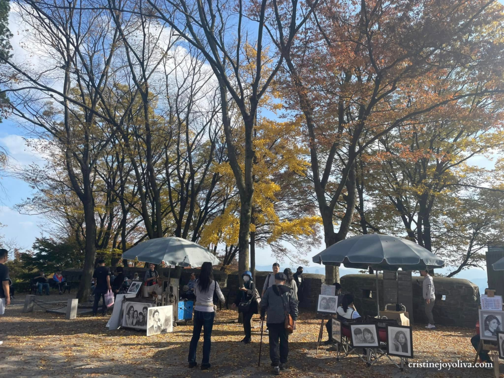 Outdoor art stalls with portrait artists under umbrellas, surrounded by tall ginkgo trees with yellow autumn leaves at Namsan Park, Seoul.