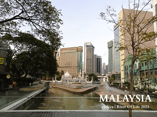 River of Life in Kuala Lumpur, Malaysia, with Masjid Jamek Sultan Abdul Samad mosque, white domes and minarets, surrounded by modern skyscrapers and greenery.