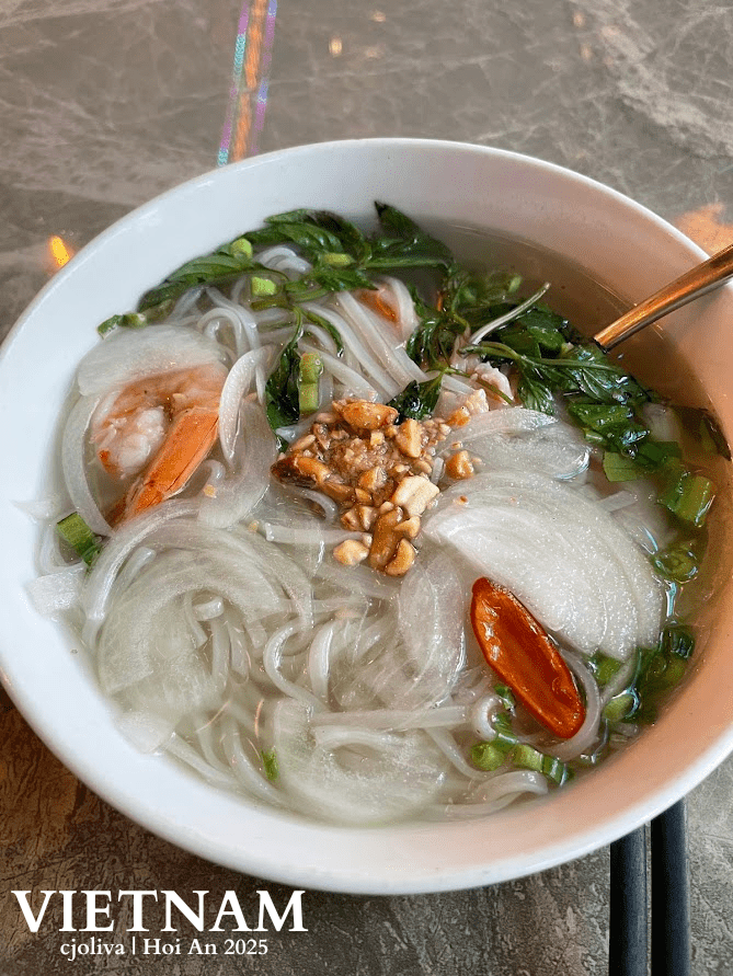 Bowl of Vietnamese prawn noodle soup at Riverside Hoi An, with rice noodles, shrimp, sliced onions, green onions, herbs, crushed peanuts, and red chili pepper in a clear broth, served with a spoon and chopsticks on a marble table.