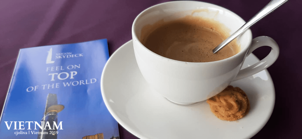 White cup of coffee with cookie on saucer, metal spoon inside, placed beside a blue brochure titled “Saigon Skydeck – Feel on Top of the World,” with an image of a tall building, on a dark purple surface.