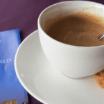 White cup of coffee with cookie on saucer, metal spoon inside, placed beside a blue brochure titled “Saigon Skydeck – Feel on Top of the World,” with an image of a tall building, on a dark purple surface.
