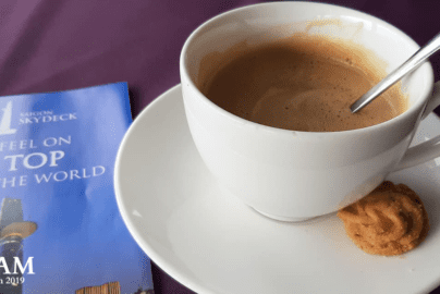 White cup of coffee with cookie on saucer, metal spoon inside, placed beside a blue brochure titled “Saigon Skydeck – Feel on Top of the World,” with an image of a tall building, on a dark purple surface.