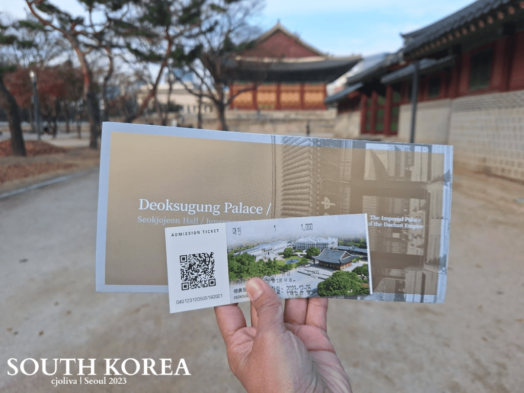 Hand holding admission ticket and brochure for Deoksugung Palace in Seoul, South Korea, with palace architecture and tree-lined path in background.