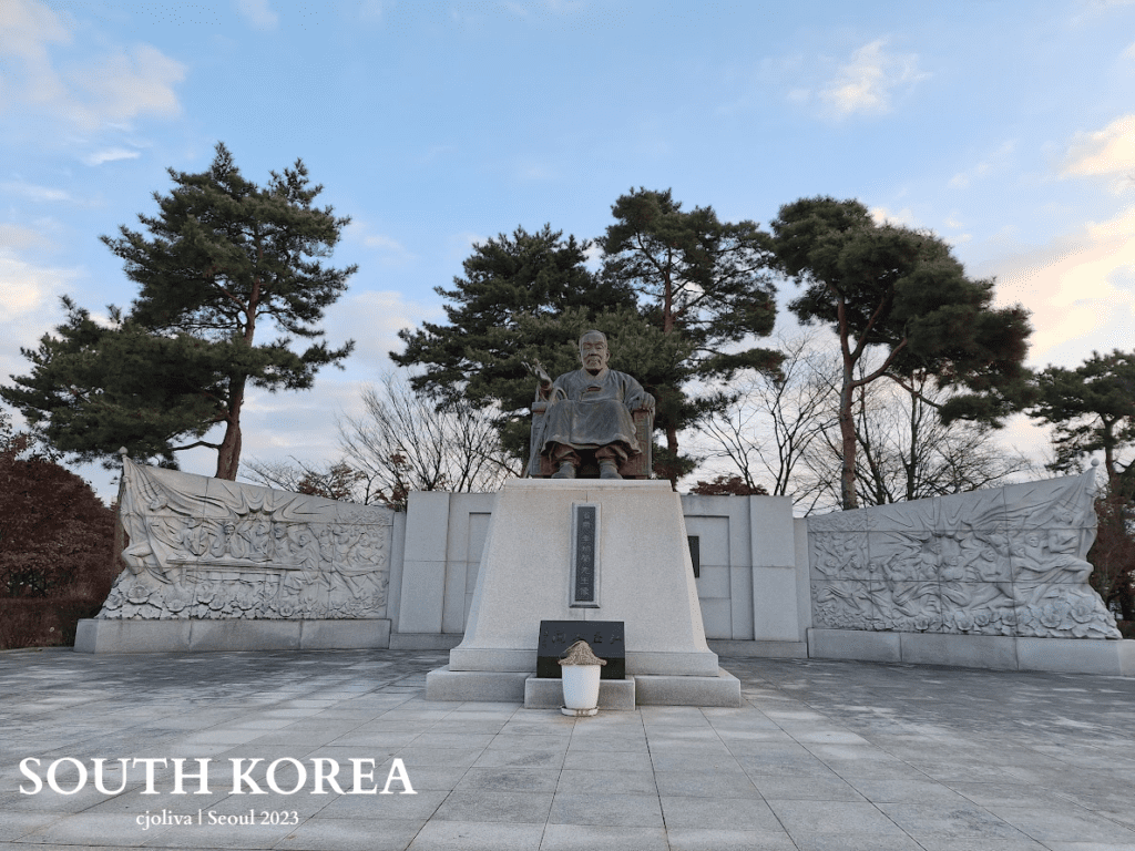 Statue of a seated figure on a pedestal with Korean inscriptions, flanked by stone reliefs and pine trees in a landscaped monument area in Seoul, South Korea.