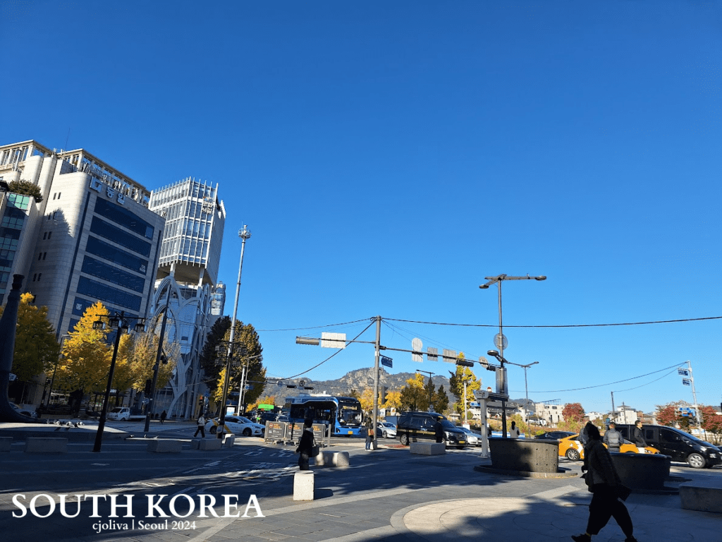 Busy Seoul street with glass skyscrapers, vehicles, pedestrians, and autumn foliage under a clear blue sky.