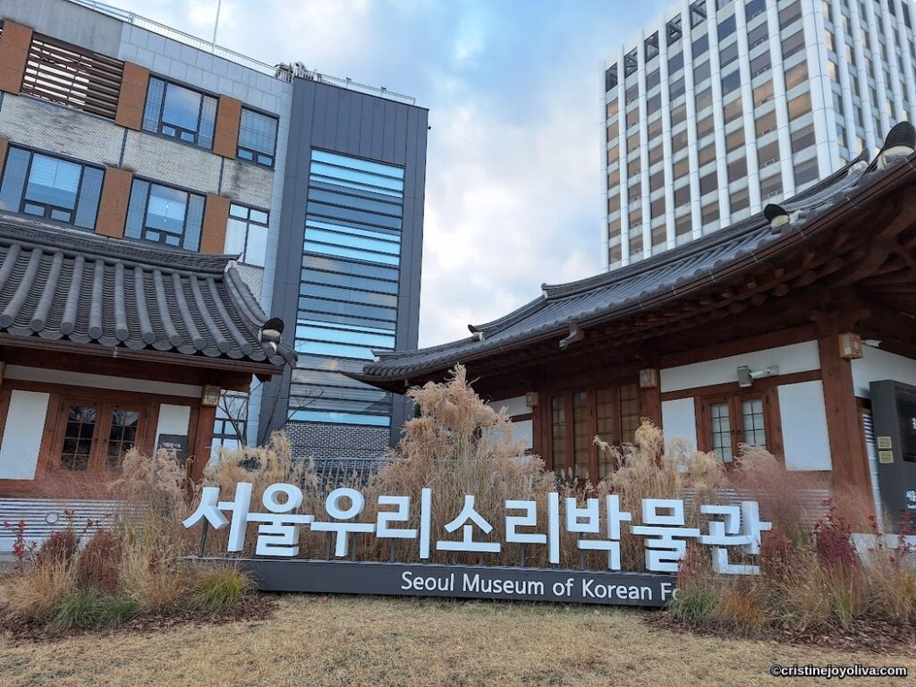 Exterior of the Seoul Museum of Korean Folk Music with traditional tiled roof and wooden frames, set against modern high-rise buildings.