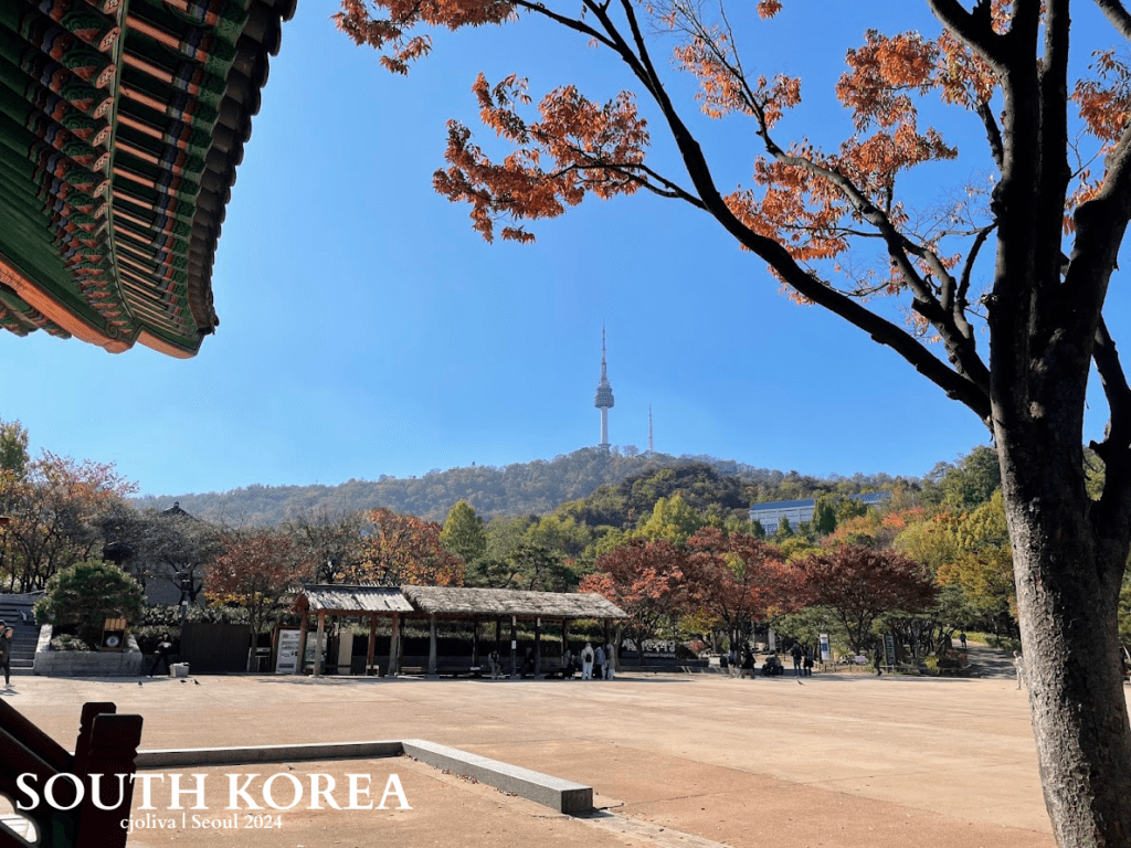 Traditional Korean courtyard with autumn foliage and Namsan Seoul Tower rising above a forested hill in Seoul, South Korea.
