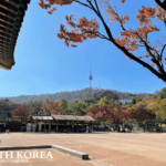 Traditional Korean courtyard with autumn foliage and Namsan Seoul Tower rising above a forested hill in Seoul, South Korea.
