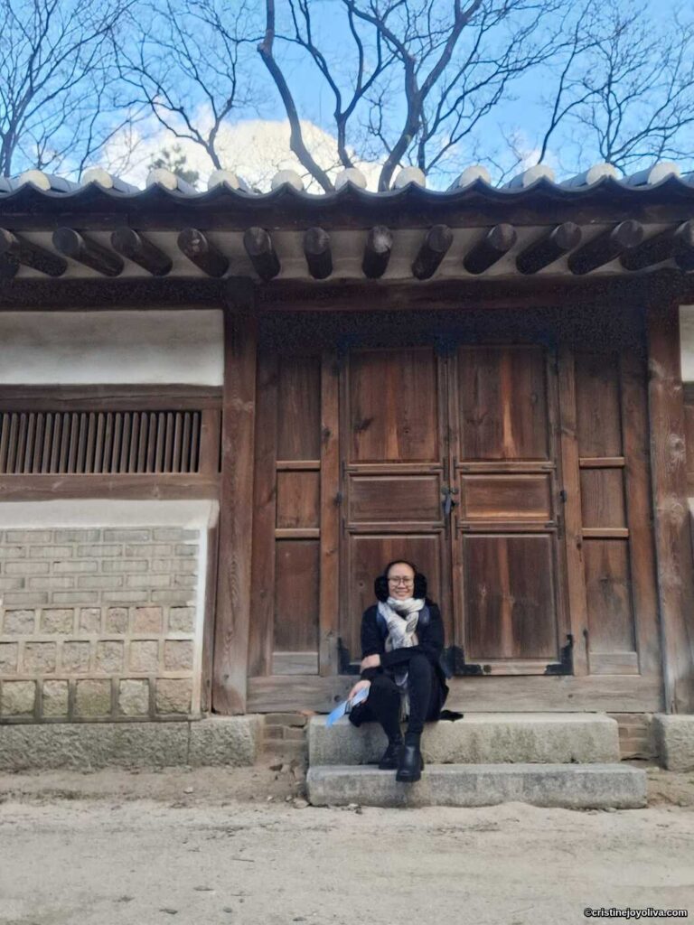 Person sitting on stone steps in front of a traditional wooden hanok building with tiled roof, brick and plaster walls, and bare tree branches under a clear blue sky in Seoul, South Korea.