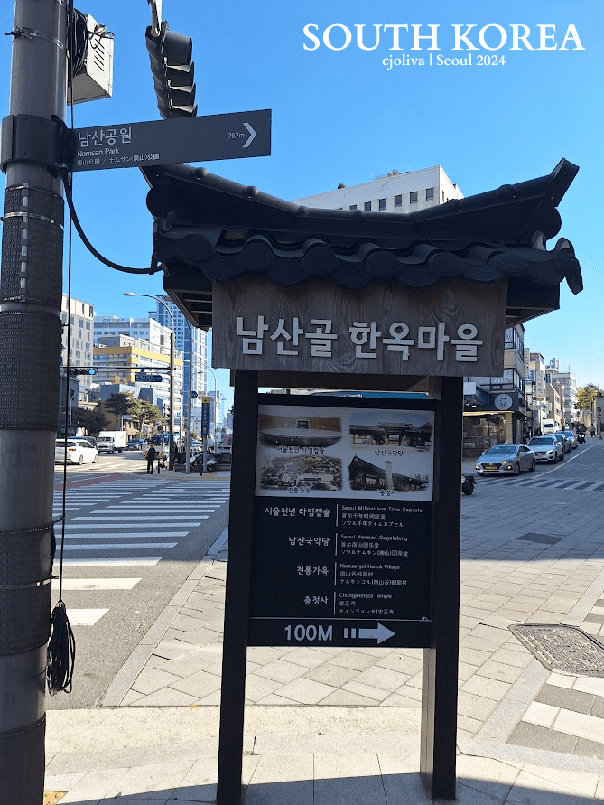 Traditional wooden signboard with tiled roof in Seoul, South Korea, showing directions to Namsangol Hanok Village and nearby attractions in Korean and English.