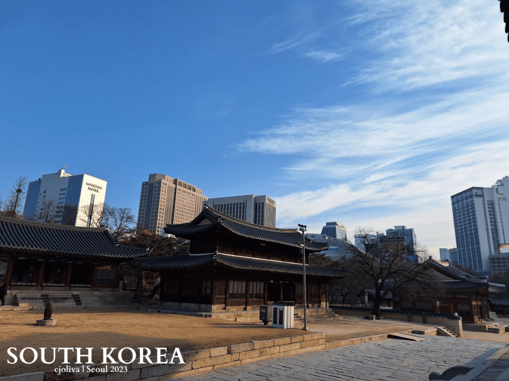 Traditional Korean palace courtyard with wooden buildings and tiled roofs in Seoul, South Korea, contrasted against modern skyscrapers, including the Koreana Hotel.