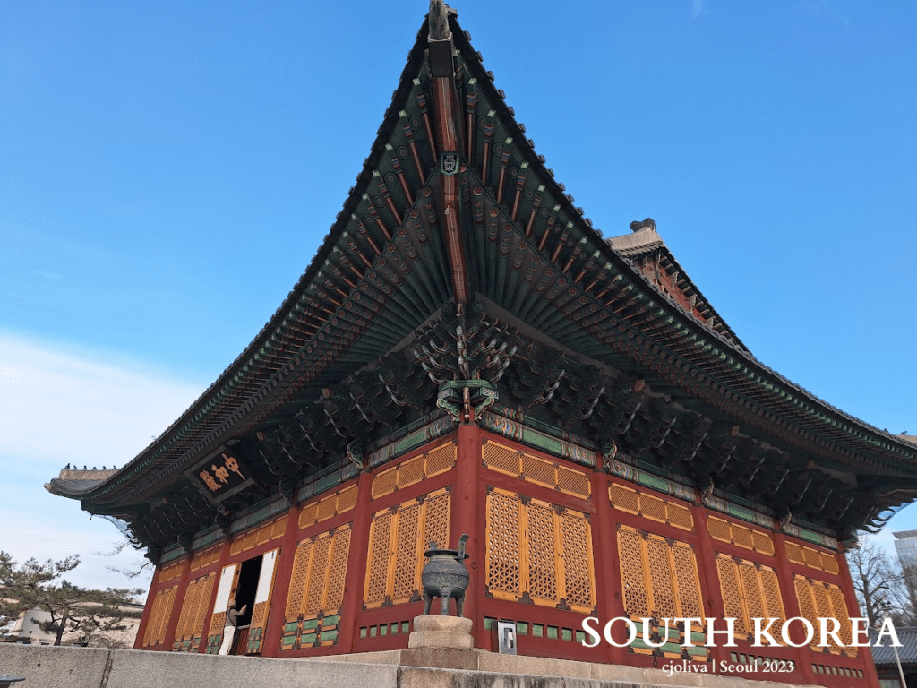 Traditional Korean palace building in Seoul with a colorful decorated roof, red and yellow lattice windows, a stone platform, and a large decorative urn in front.
