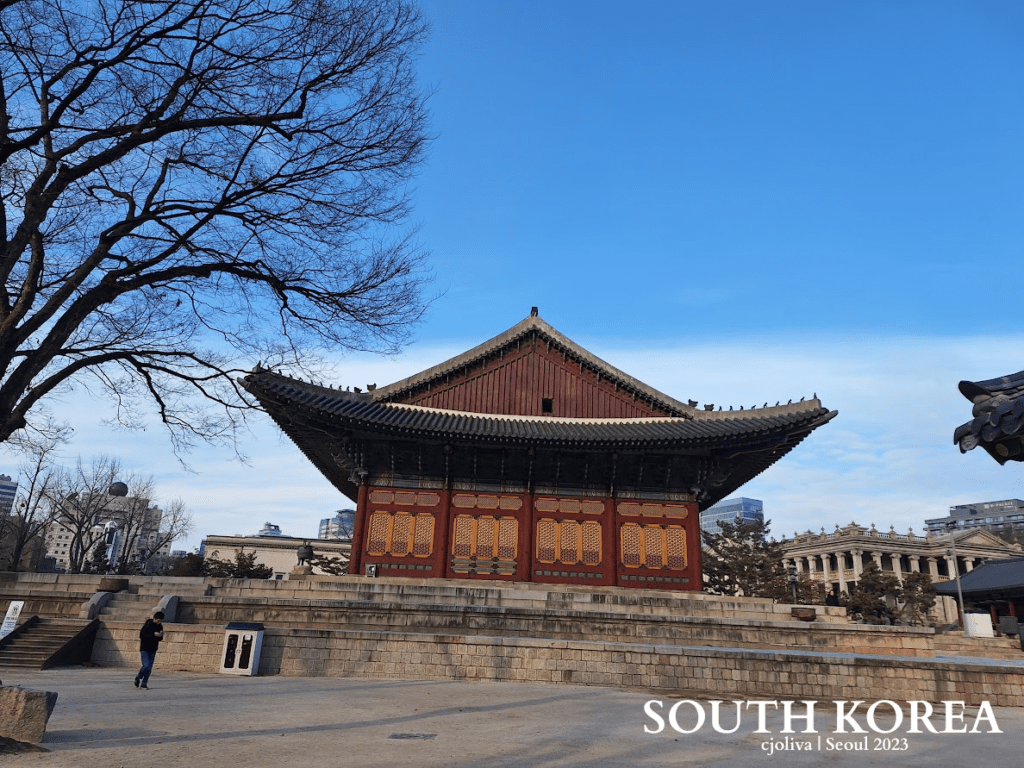 Traditional Korean palace building in Seoul with a curved tiled roof, colorful wooden panels, and a stone platform, framed by a large tree and modern buildings in the background.