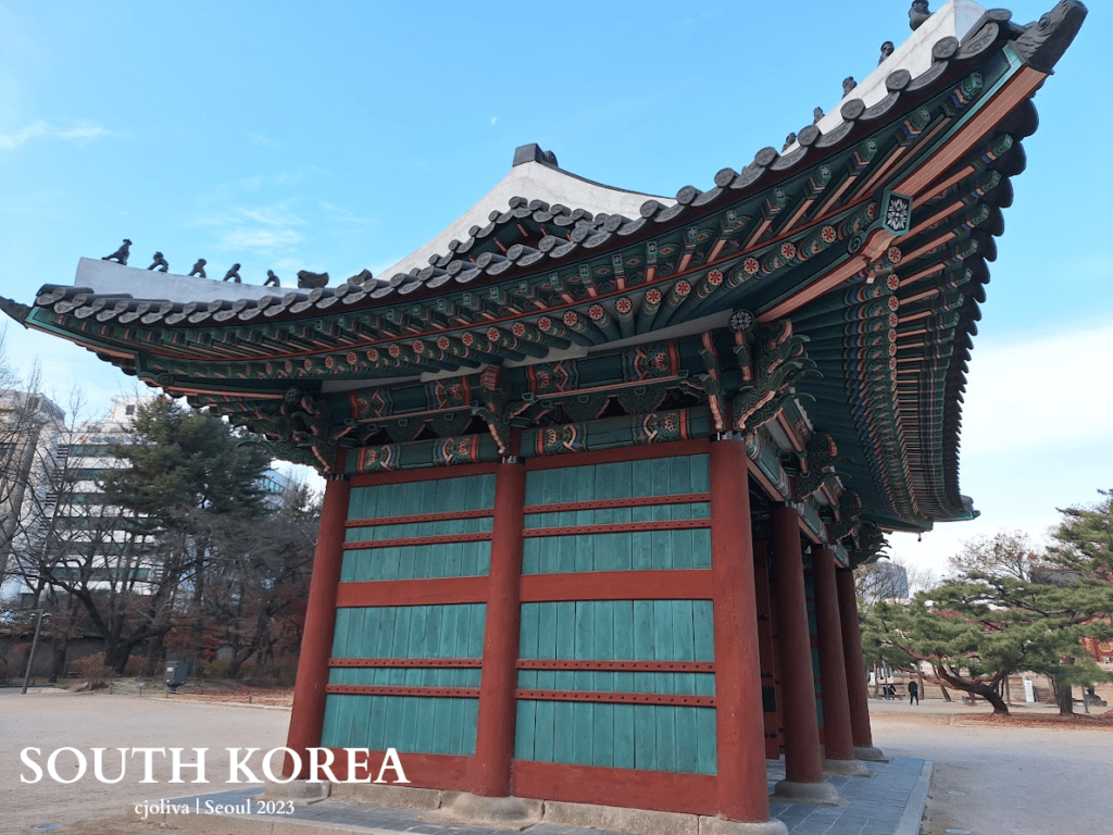 Ornate traditional Korean gate pavilion in Seoul with colorful wooden beams, tiled roof, decorative figurines, and stone platform surrounded by trees and modern buildings.