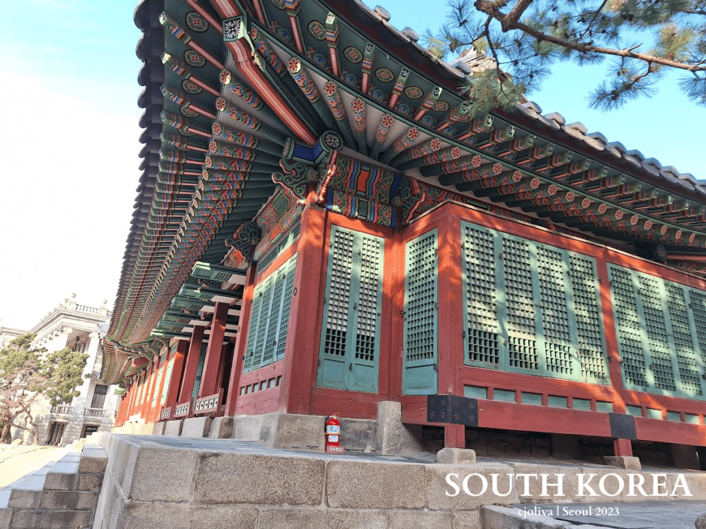 Traditional Korean palace building in Seoul with red wooden pillars, green lattice windows, and decorative painted eaves on a raised stone foundation.