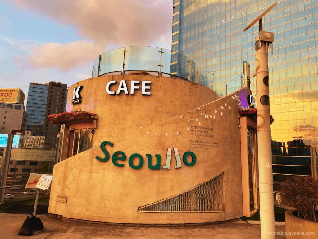 The exterior of K-Cafe on the Seoullo 7017 elevated walkway during sunset, with the golden light reflecting off a modern glass skyscraper in the background.